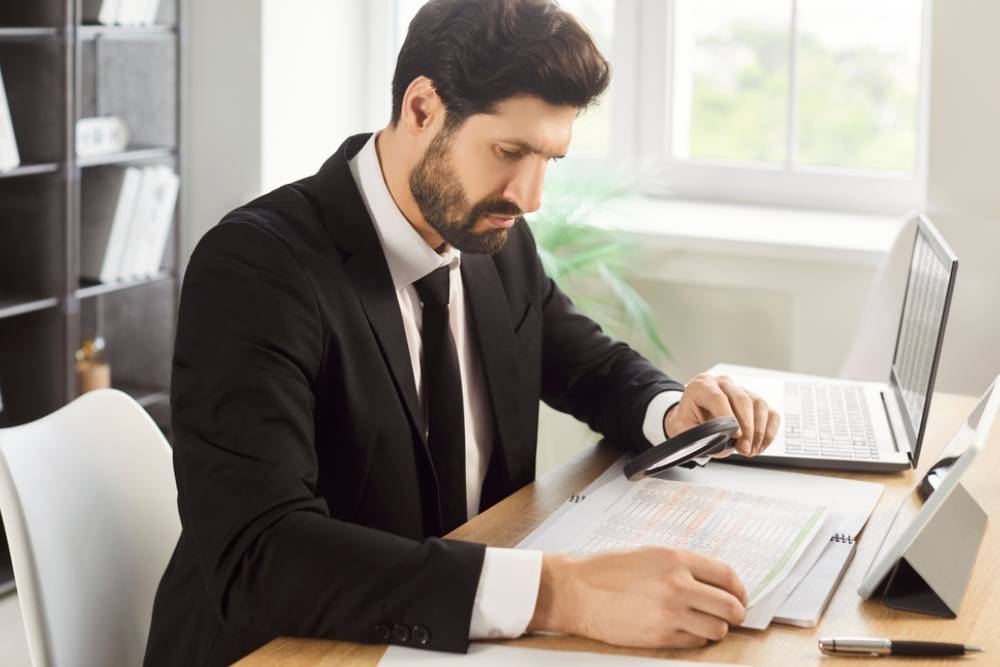 Analysis of business documents by busy businessman with magnifying glass in office